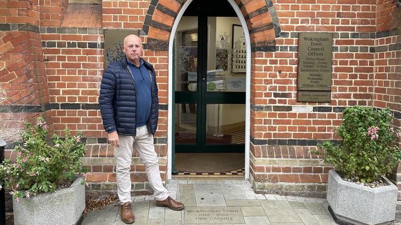 A man wearing a navy puffa jacket, navy polo shirt, cream trousers and brown shoes stands by an open door in a red brick building.