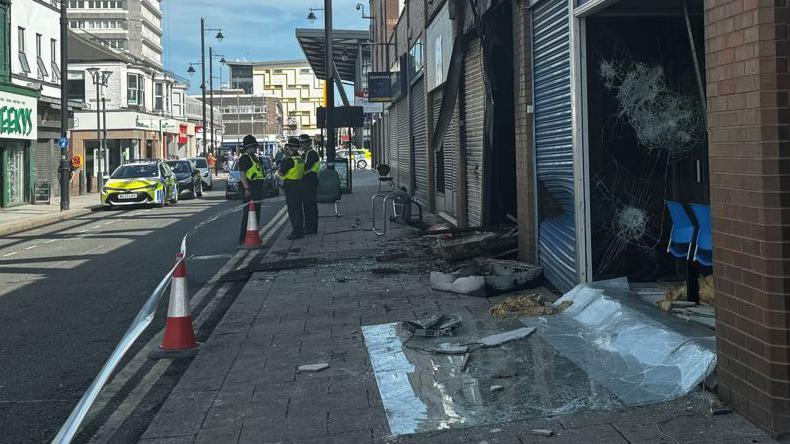 Broken window on the ground in front of a police office. Police officers are standing further up the street and debris from the burned out unit next door spills out on to the pavement.