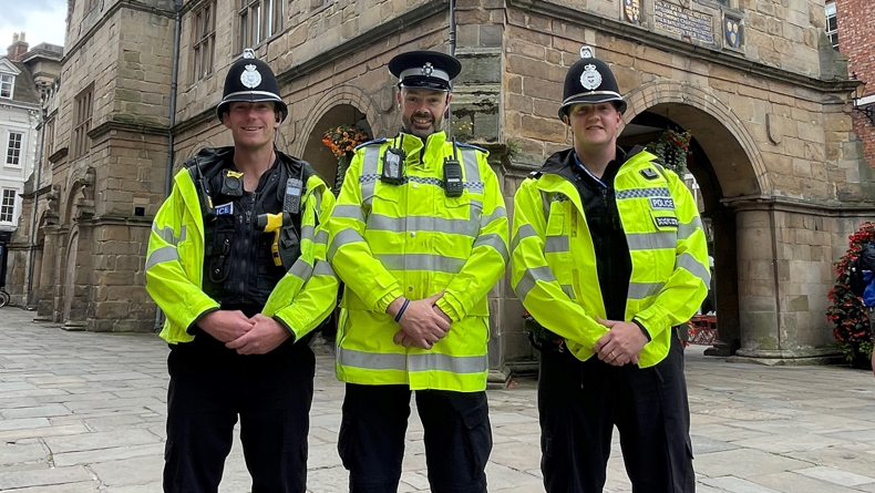 Three police officers in high-vis jackets stood in a market square smile at the camera