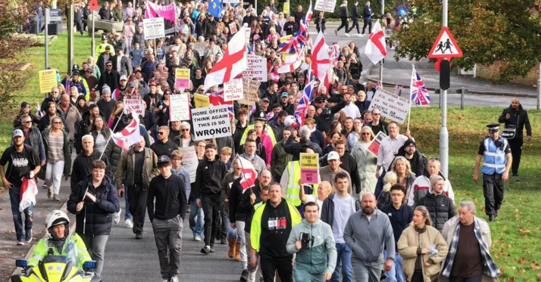 A number of people at a protest in Crowborough, East Sussex, on 8 November. Demonstrators are resisting plans to house asylum seekers at the former army camp in the town. A number of England and union jack flags can be seen in the image, alongside a few placards.