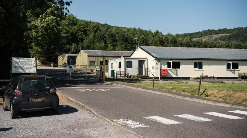 A section of the former Yeo Valley Farm site in Cheddar. It shows an entrance driveway with a zebra crossing and a metal gate securing the site. There are long one-storey buildings with corrugated metal roofs. In the distance there are fields and mature trees.