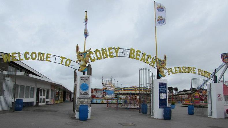 Entrance to the amusement park on a grey day with gates open and lettering above saying welcome to Coney Beach Pleasure Park. Attractions are visible behind including a caroussel.