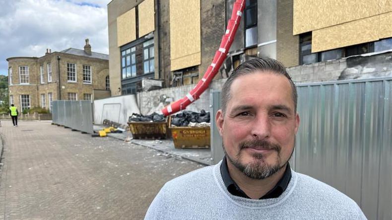 Kent County Councillor James DeFriend stands in front of the fire ravaged Conqueror House on Crabble Hill, Dover. A rubble chute, temporary fencing, boarded up windows and smoke damage cover the apartment building