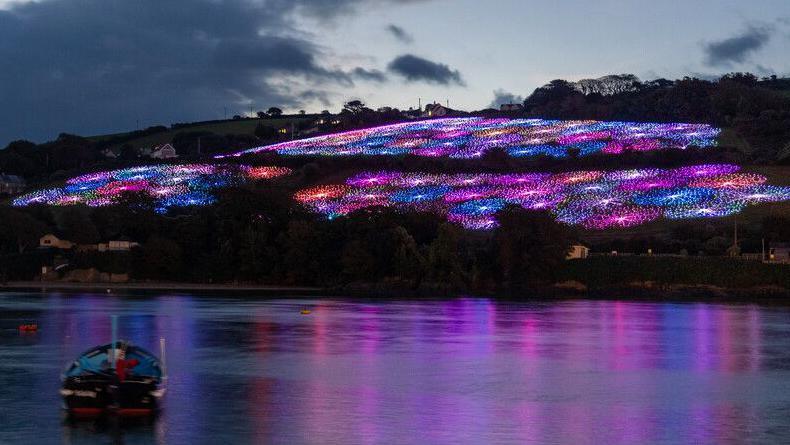 Fibre optic lights illuminate a field overlooking Salcombe harbour. The lights are in patches of colour including blue, pink, purple, orange and white. There is a pink glow on the water from the lights and a small fishing boat in the foreground.