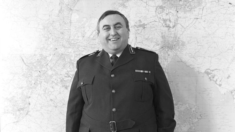Black and white photo of Alan McQuillan wears a dark blazer embellished with pins. He has short, dark hair and is smiling in front of a map of the world.