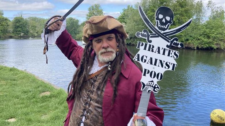 Darren poses in his pirate clothing holding a cutlass over his head and a sign saying "Pirates against Poo", which has a skull and crossed cutlasses at the top. He wears a maroon jacket, laced white shirt and brown tunic, and stands by the side of a river.