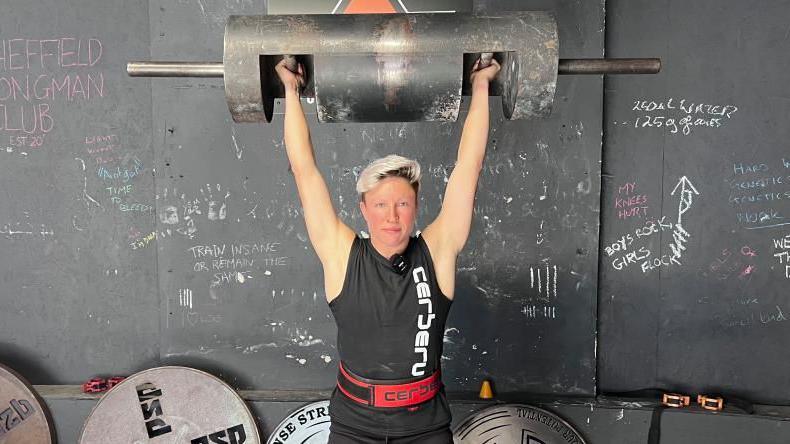 Laura Hollywood stands in a gym holding a large metal log bar overhead with both arms extended. She is wearing a black sleeveless top with white vertical text and a red lifting belt. Behind her is a dark wall with chalk writing and several large weight plates stacked on the floor.