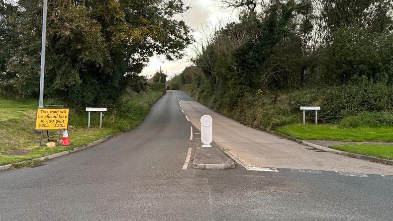 The junction at Cooil Road. A yellow and black sign sits next to the road and there are green grassy patches and trees at either side of the empty carriageway.