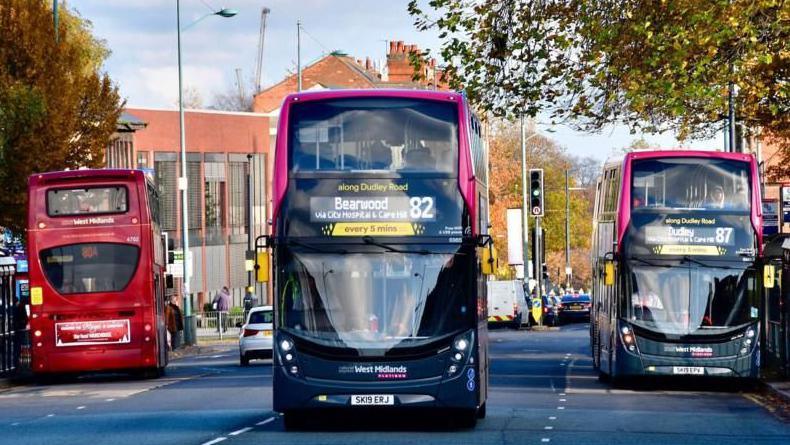 A three-lane road with three buses on it. A red bus is at a bus stop facing away from the camera. Another bus, with Dudley marked as its destination, is at a bus stop on the other side of the road facing the camera. Between them, a bus heading for Bearwood is driving towards the camera.