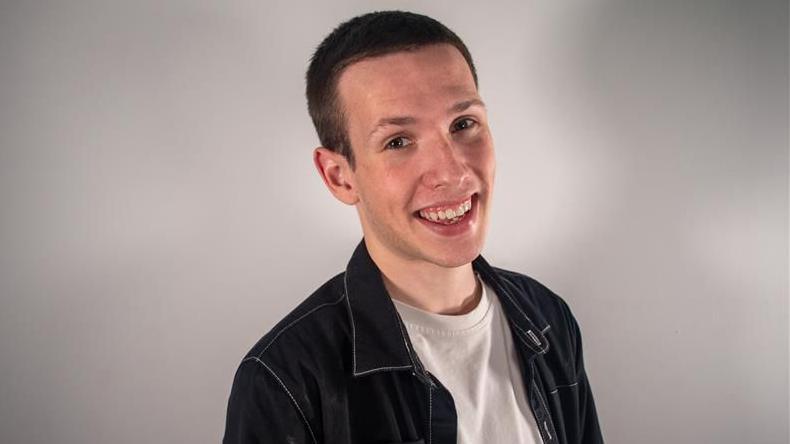 Matt Hallsworth, in a white t-shirt and black jacket,  looking directly into the camera. He is smiling and has short brown hair.