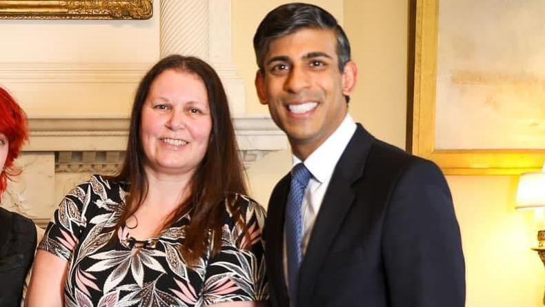 Chrissie Lowery and Rishi Sunak. Chrissie has long dark hair and is wearing a black top with a leaf pattern on it. Rishi Sunak is standing next to her. He is wearing a white shirt, blue tie and a black suit jacket. They are both looking at the camera and smiling. 