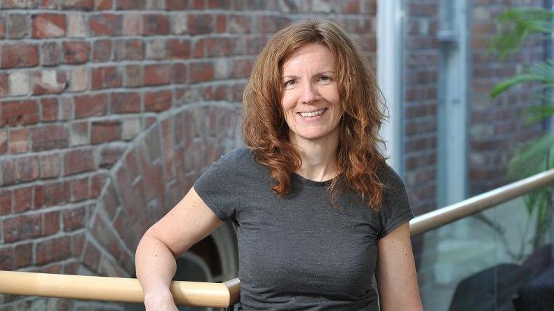 Karen Winter has long wavy ginger hair and is wearing a grey t-shirt. She is standing by a red brick wall and glass wall on an inside balcony. 