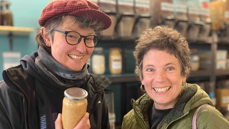Kimberley Moore, wearing glasses, a black jacket and red hat, holding up a jar of peanut butter, with Tara Sampy standing next to her in a green jacket