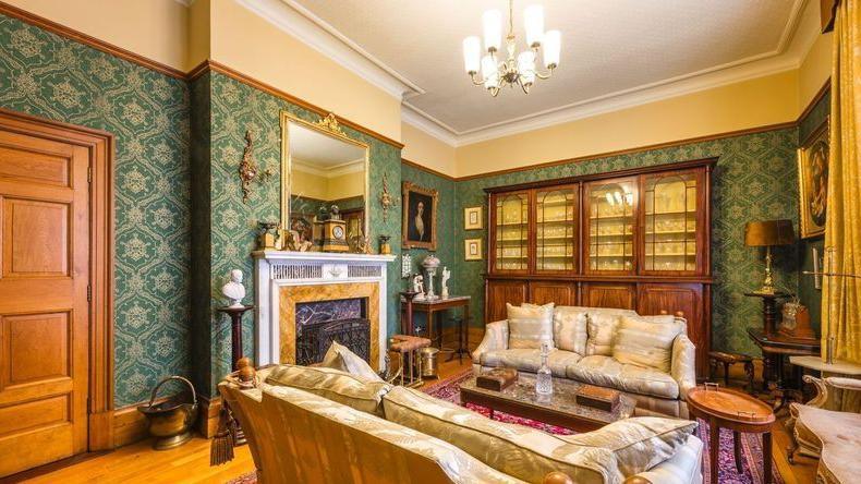 A lounge in the Bishop's House in Newry.  The room has ornate, dark green wallpaper and a marble fireplace.  Two sofas face opposite each other, separated by a marble-topped coffee table.  A large glazed display cabinet lines the back wall. 