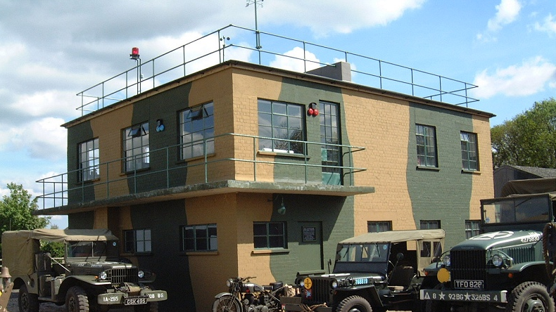 Twinwood Control Tower pictured in present day, in colour. It is a square 2-storey building painted in military colours, khaki beige and green. There is fencing around the top and first floor balcony, and several military jeeps parked in front. A cloudy blue sky overhead.