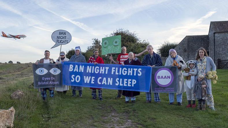 Members of the Bristol Airport Action Network standing on Felton Common on a cloudy day. They are all wearing pyjamas and dressing gown and are carrying signs which say "ban night flights"," eyes on Bristol airport", "ban night flights so we can sleep". There is an EasyJet aircraft flying in the background.