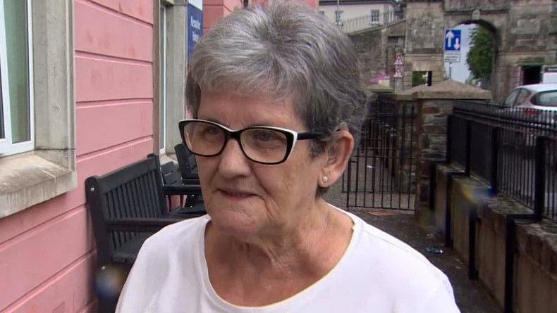 Catherine Coyle has black and white rimmed glasses and is wearing a white T-shirt. She and is standing outside a pink building with a row of black benches behind her and gates.