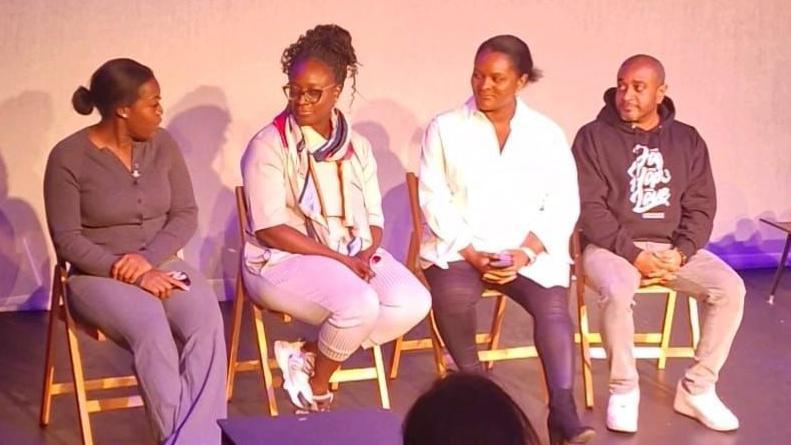 Three women and a man sitting on chairs on a stage.
They are on a panel and sitting in front of a audience