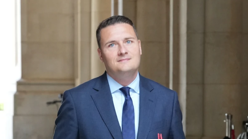 Wes Streeting looks directly at the camera as he walks outside. He is wearing a blue suit and blue tie, and a stone building is behind him. 
