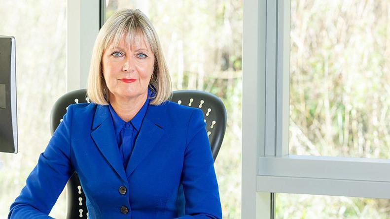 Joy Allen is sitting at her desk. She has straight blonde hair, cut in a bob. Her eyes are blue and she is wearing a blue shirt and jacket.
