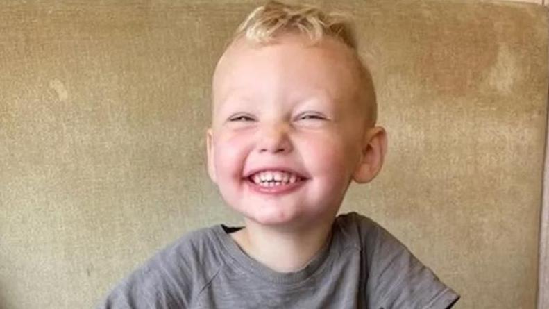 Boy with blonde curly hair on top and shaved at sides grins into camera. He's wearing a grey t-shirt