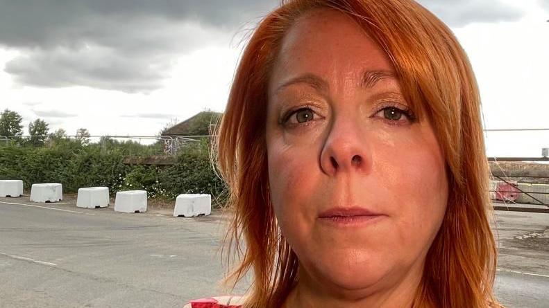 Julie Ford stands in front of the site Kempston Hardwick which has been bought by Universal Destinations and Experiences. She looks directly at the camera. She has red shoulder lenght hair and wears a red and white patterned top.