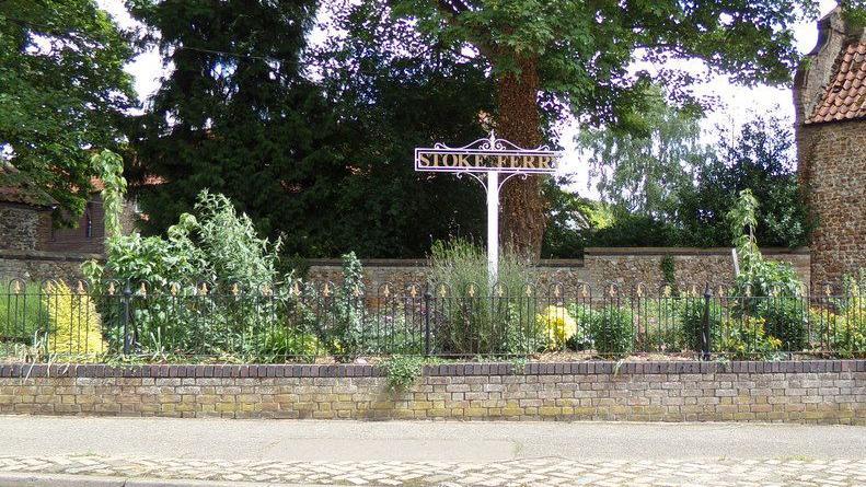 Stoke Ferry's village sign. The ornate sign on a white pole stands in a small fenced-off garden. There is a flint cottage nearby.