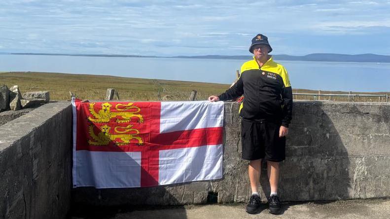Kevin Adams is smiling as he stands next to the flag of Sark by the coast, with the sea in the background. He is wearing the yellow and black kit worn by the Sark team.