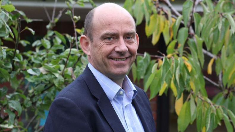 Toby is wearing a light blue shirt with dark blue jacket. He is standing in a garden with plants and shrubs behind him. 