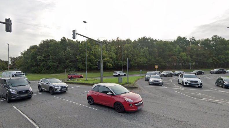 Traffic entering onto a roundabout. A row of cars has stopped at traffic lights on the roundabout. There are overhead traffic lights, and trees are visible alongside the road in the background.