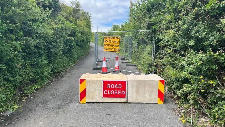 A country road is closed with concreate barriers with a red 'road closed' sign on it. There is also metal cage fencing behind the barrier with a yellow sign that says 'Pedestrian access only, please keep away from cliff edge'.
