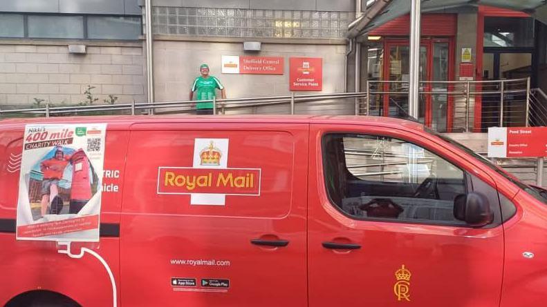 A royal mail van parked outside its building. Nikki is stood against the building, behind the red van. She is smiling at the camera. On the back of the van is a poster which has a picture of Nikki and '400 miles' above it.