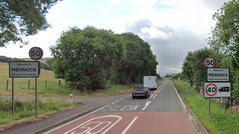 A black car and a white caravan drive along a tree-lined road with signs either side saying Welcome to Mennock