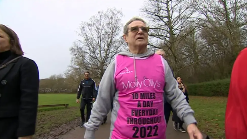 A man with short grey hair walks through a park, accompanied by a number of other people. He is wearing a grey hoodie and a pink tabard which reads: "Molly Ollys. 10 miles a day everyday throughout 2022."