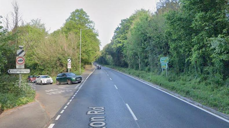 The junction of a rural A-road with a side road. A queue of cars are waiting to join the A-road. Trees line both routes. Traffic signs showing the national speed limit are visible.