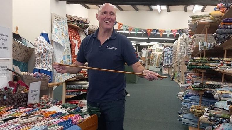 Bryan Machin wearing a blue polo shirt and standing in the haberdashery store. He is holding a metre stick