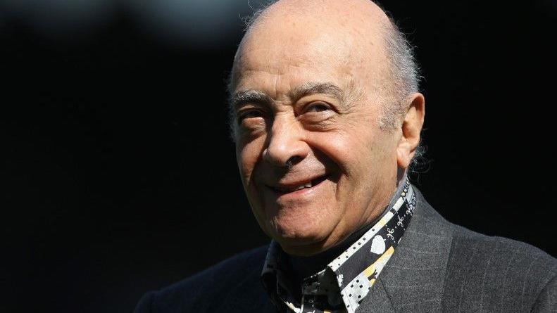 A close-up of Mohamed Al Fayed's head and shoulders. He is smiling at the camera and is wearing a dark grey suit with stripes, and a patterned shirt.