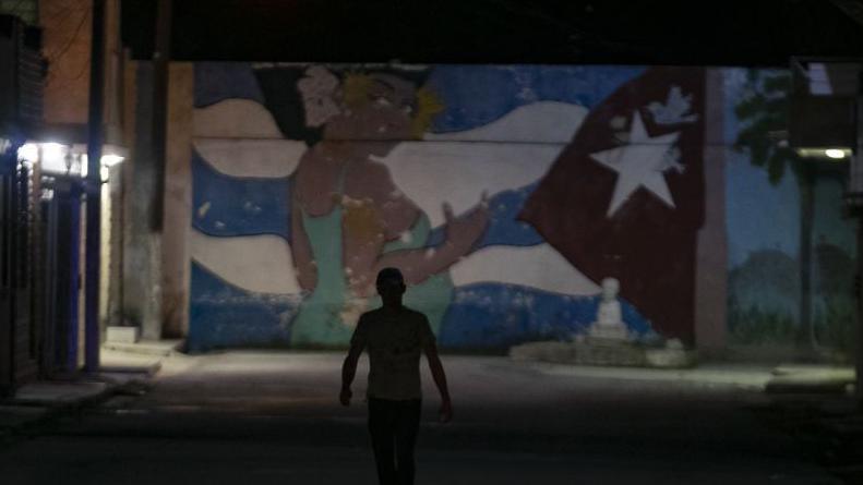 A pedestrian walks down a street during a rolling blackout in Havana. He's in shadow but a wall mural is highlighted ahead of him. The mural features a woman in a green dress and a Cuba flag motif.