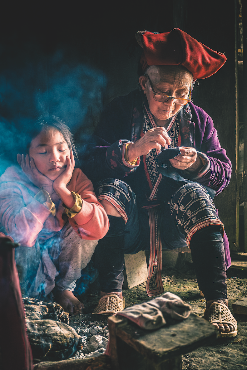 a girl sits with her head in her hands appearing to be asleep while smoke trails upwards, she sits next to her great grandmother who is working on her embroidery