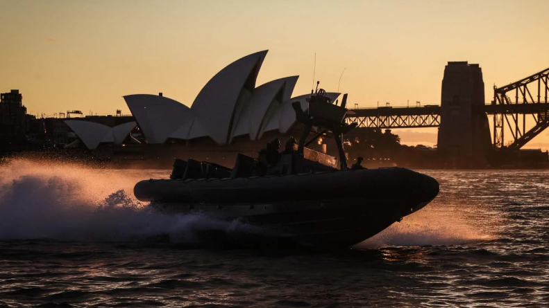 A picture of a boat conducting the operation in front of the Sydney Operah House. It is a small boat with a bridge in the background of the image too.