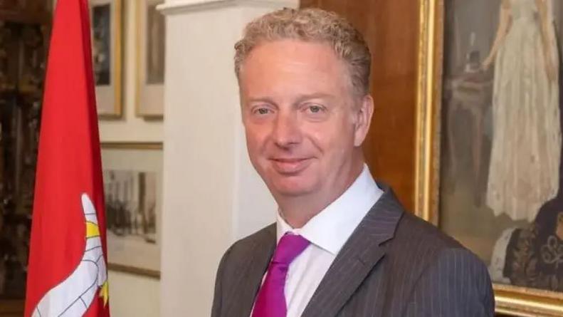 Alex Allinson, a man which short light coloured hair. He is wearing a grey suit with a white shirt and purple tie and is standing next to a Manx flag in an office.