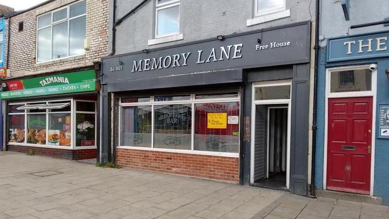 Memory Lane's entrance. It is black door with three windows to the left. There is a writing above the door reading est. 2021 Memory Lane Free House. There is a takeaway next to it on the left.