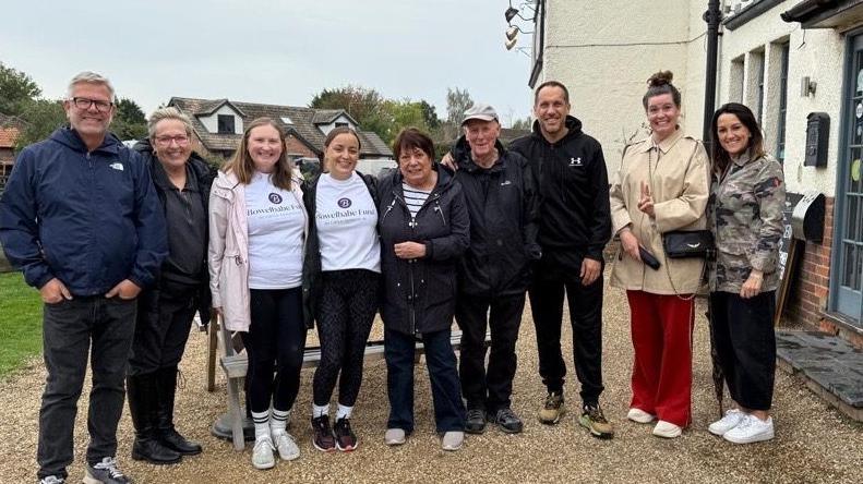 Nine people are lined up outside a pub. They are all looking to the camera and smiling. They are stood on a gravelly area and have trainers or walking boots on. There is a cream coloured building behind them to the right. 