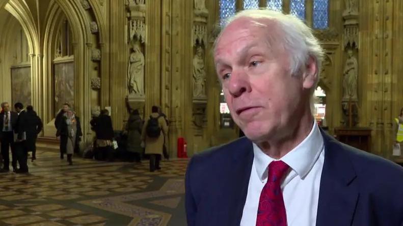 MP Si Nic Dakin in the Palace of Westminster. He has short white hair and wears a smart blue suit, white shirt and red tie.