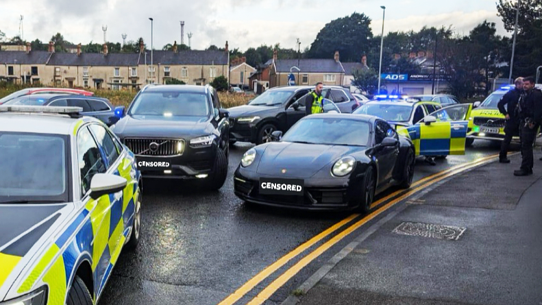 Stolen black Porsche at the side of a road surrounded by police vehicles, and with police officers standing nearby