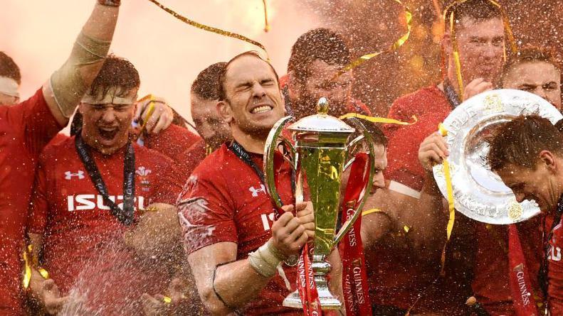 Alun Wyn Jones holding the 6 Nations trophy. He is closing his eyes and celebrating, while other players celebrate around him. 