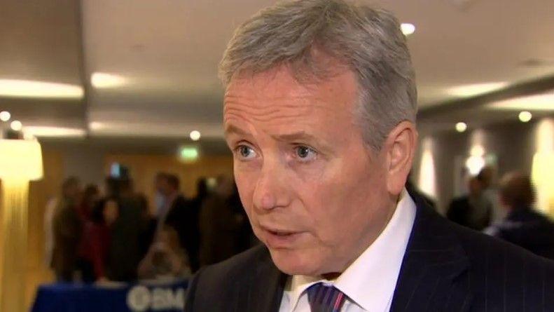 Dr Tom Black wearing a black suit, white shirt and coloured tie. He is standing with a room of people behind him. He has grey hair.