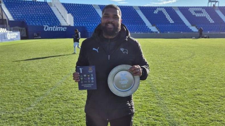 Derby County Under-12s lead coach Zeph Thomas with the trophy after his side won the Orange Veins Madrid tournament in 2025