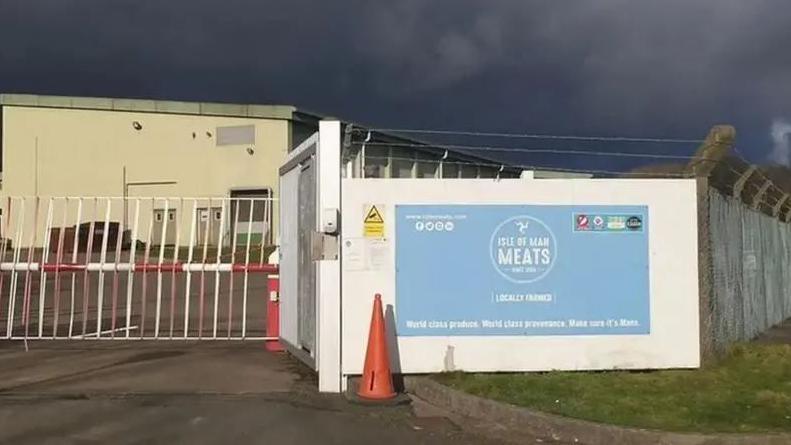 The exterior of the Meat Plant with a blue a sign that reads Isle of Man Meats on a white board, which is attached to a tall fence topped with barbed wire.