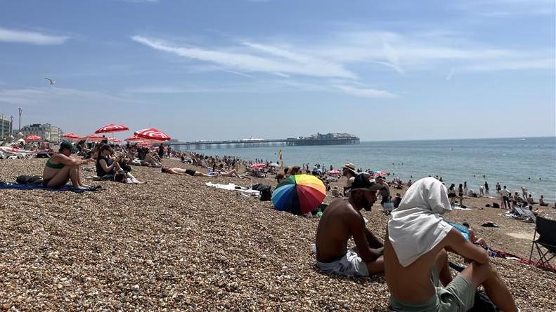 A number of people sat on the stony Brighton beach soaking up the sun. A number of umbrellas can also be seen in the image.